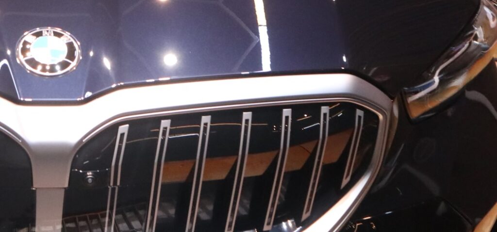 Close-up of a vehicle front showing painted panels, plastic grille, metallic trim, and a headlight made from different exterior materials.